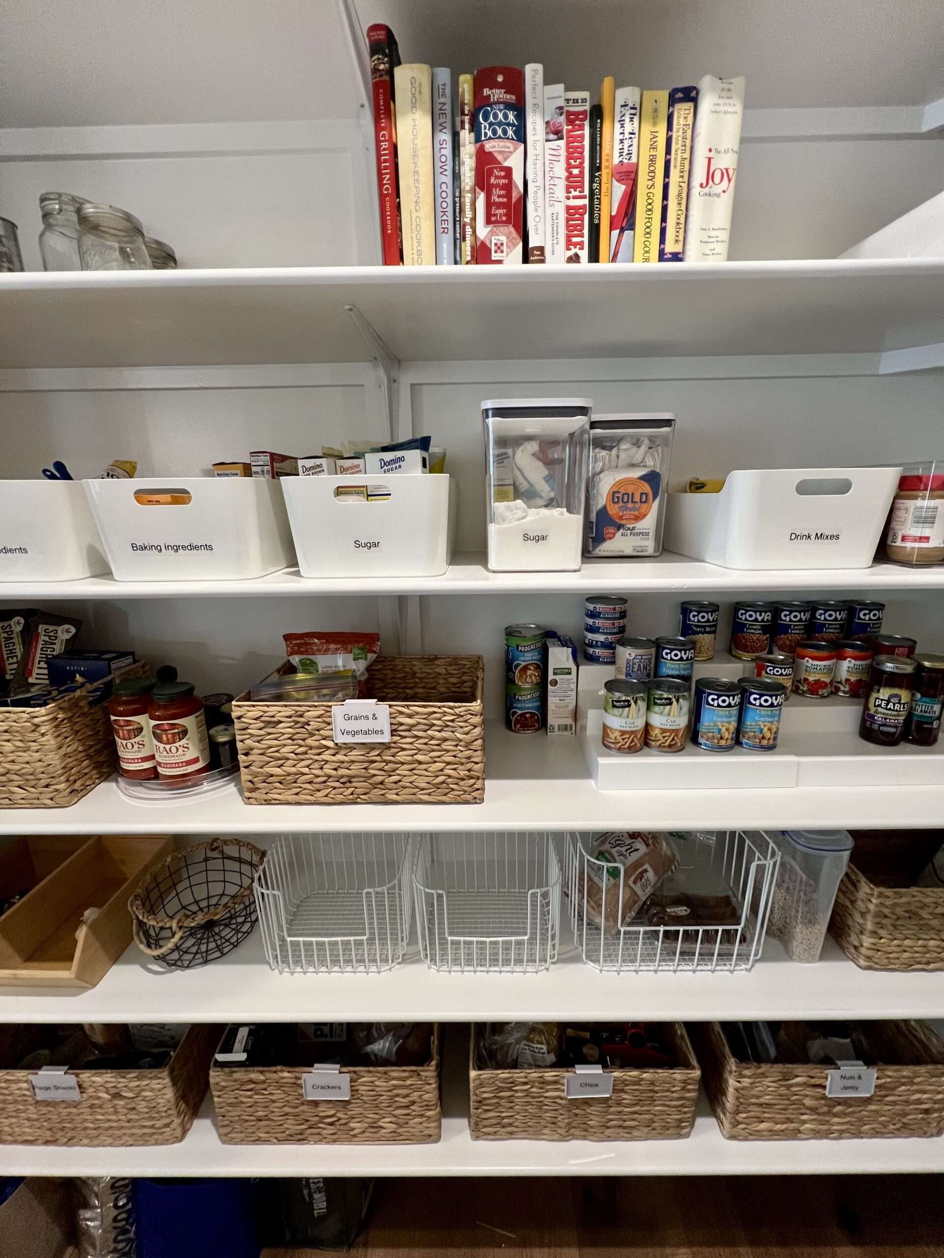 Kitchen pantry with groceries and items organized on shelves. Bins and baskets contain various food categories, and cookbooks appear on a top shelf.
