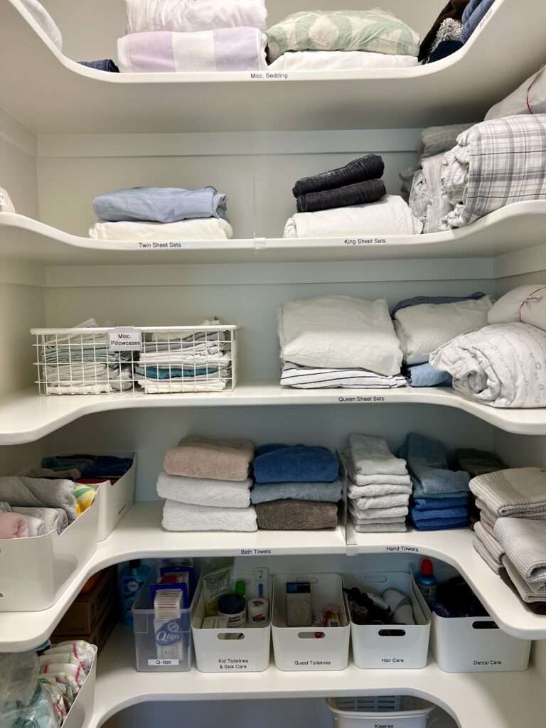 An organized linen closet. The bottom shelf includes toiletry and bath items organized into white bins. The middle and top shelves include bath and bed linens, sorted by size and type, and kept organized by acrylic shelf dividers.