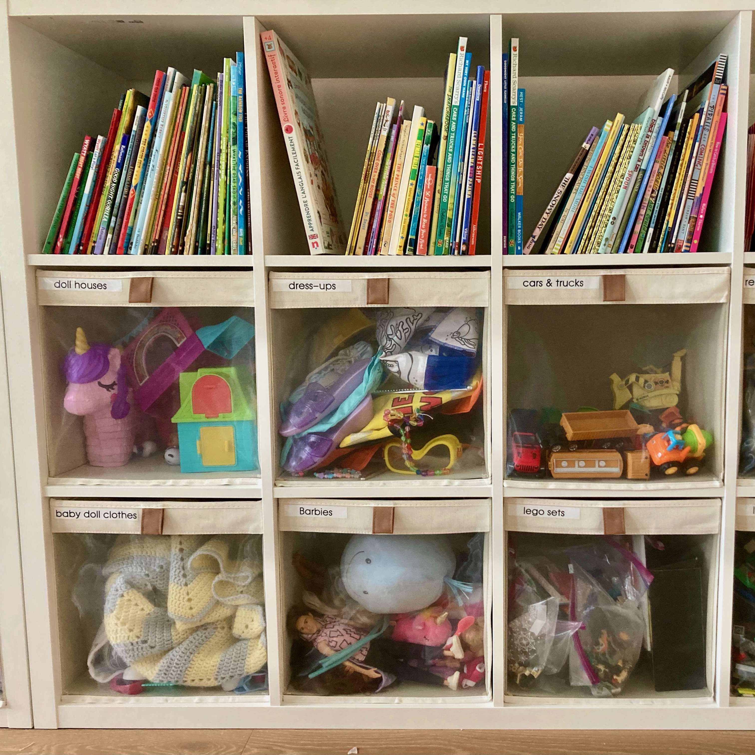 An organized playroom with toys inside an IKEA KALLAX unit. Each 13 inch bin contains a different toy category; the bins are see through on the front for easy visual access to each toy group. On the top shelf are children's books.