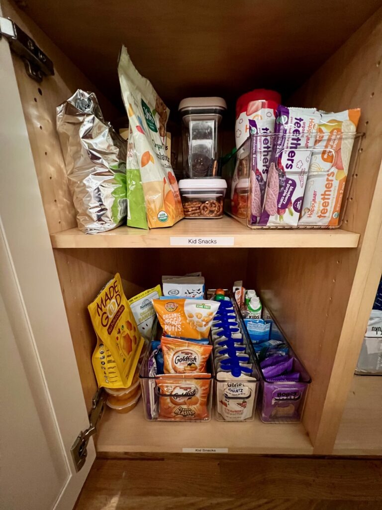 A kitchen pantry cabinet containers dedicated bins storing kid snacks. Snacks are in acrylic bins and labeled.